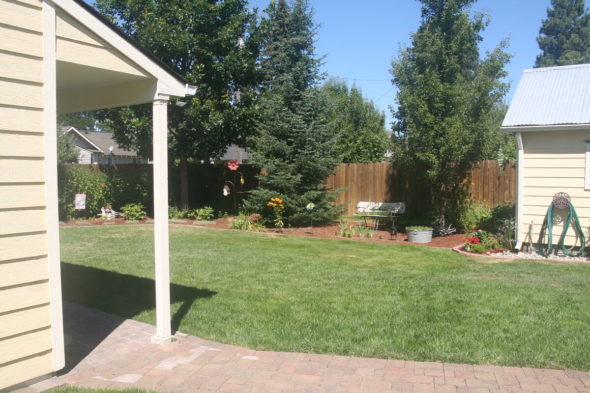 A well-maintained backyard is showcased, featuring lush green grass and a variety of trees and shrubs. A flower garden adds color, with a small seating area visible under a tree. A gravel pathway leads from the house to the inviting outdoor space.