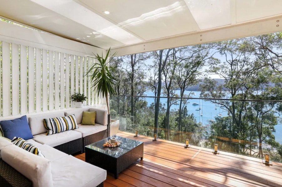 A covered deck area features a comfortable corner lounge setting with cushions and a coffee table. Glass balustrades provide unobstructed views of the water and trees, while natural light fills the space through overhead panels.