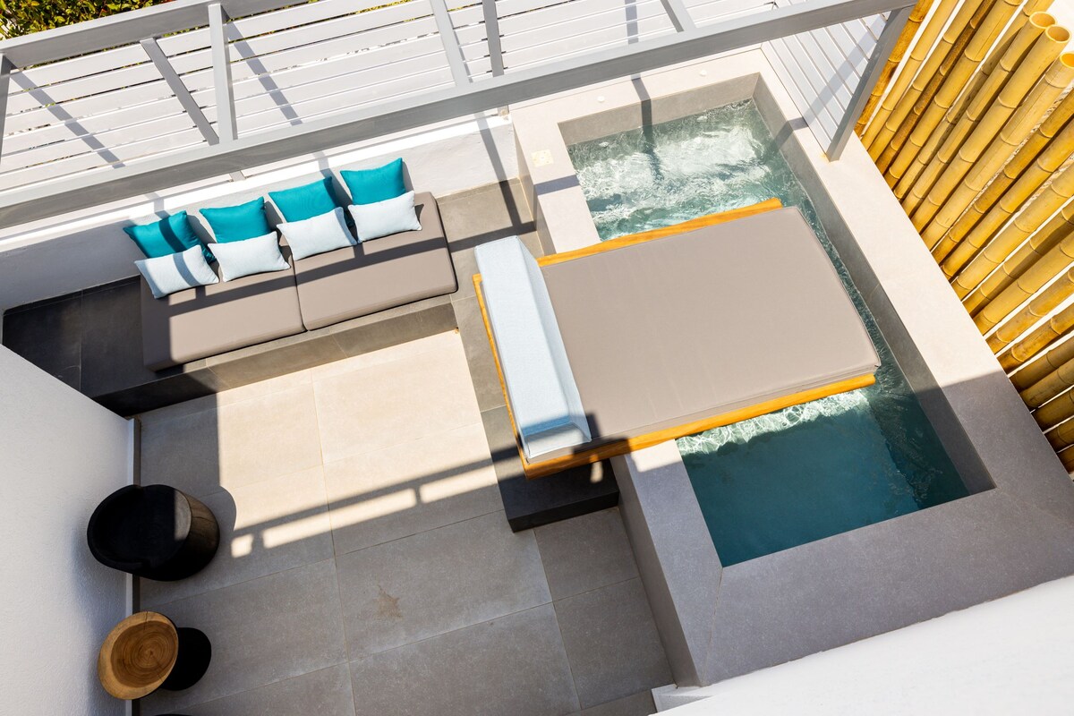 An outdoor space features a private hot tub beside a modern lounge area. Neutral stone tiles are complemented by a timber accent wall. Cushions in shades of turquoise enhance the seating, while a sleek surface extends over the hot tub for relaxation.