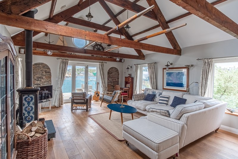 The living area features exposed wooden beams and warm wooden flooring. A spacious sectional sofa with striped upholstery is paired with a round table. Natural light floods the space through large windows, offering views of the waterfront, while a traditional wood-burning stove adds a cozy touch.