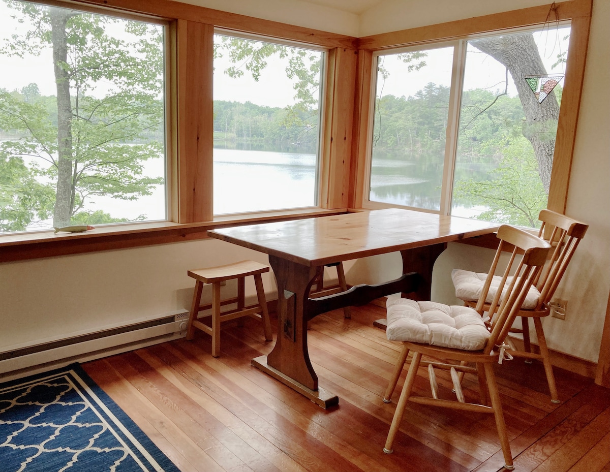 A dining area featuring a wooden table complemented by two chairs and a bench. Large windows provide expansive views of the serene lake and surrounding trees, allowing natural light to fill the space. The wooden floor adds warmth to the room.