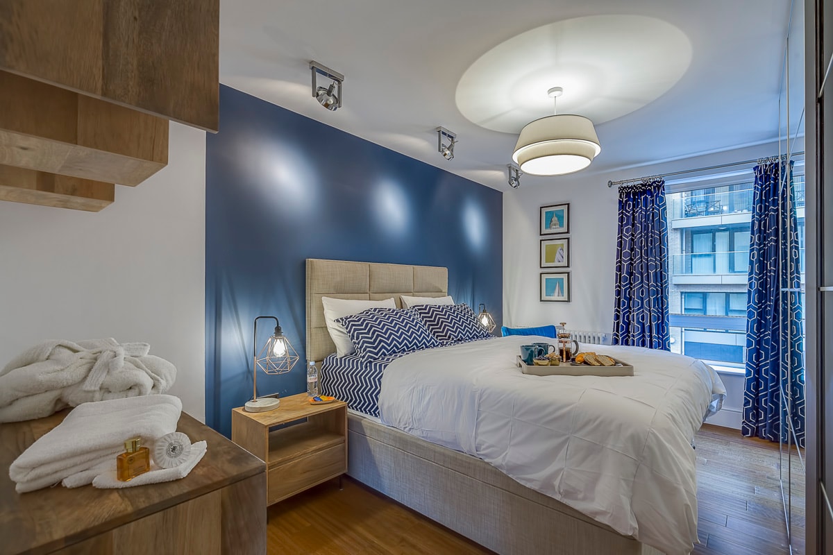 A spacious bedroom is presented, featuring a comfortable bed with crisp white bedding and a blue accent wall. Decorative throw pillows enhance the layout. Bedside tables with stylish lamps provide lighting. Large windows dressed with patterned curtains allow natural light to fill the room.