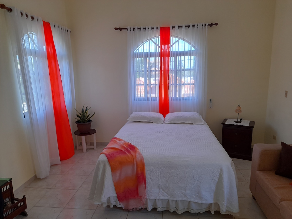 A bright and airy bedroom features a spacious bed dressed in white linens. Natural light filters through large windows adorned with sheer and vibrant red curtains. A potted plant adds a touch of greenery, alongside a nightstand with a lamp and a cozy sofa.