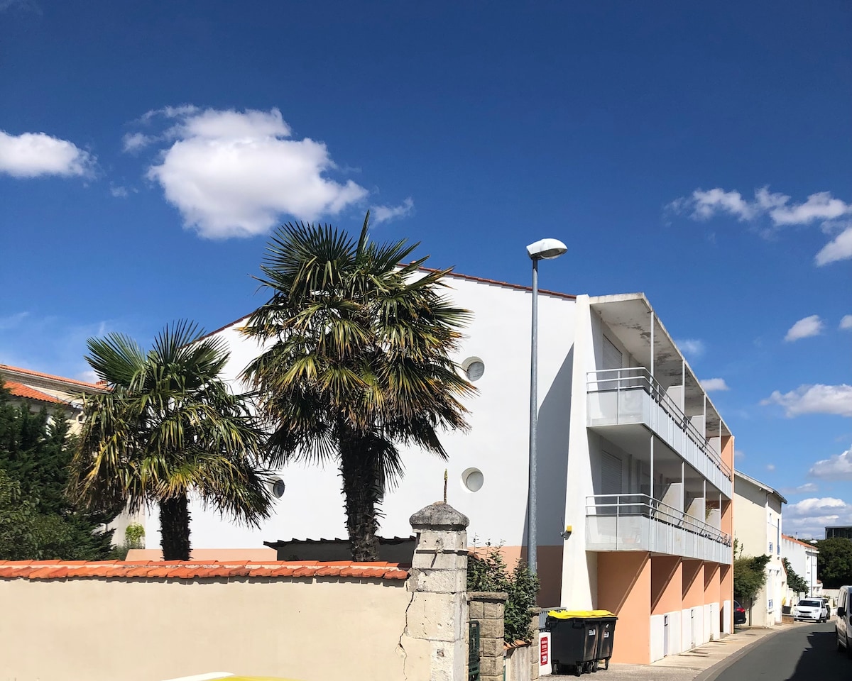 The exterior of a modern three-story building is depicted, featuring a clean design with white and peach-colored walls. Surrounding palm trees add greenery to the scene. A pathway leads to the entrance, while a large window and balcony are visible on the upper level.