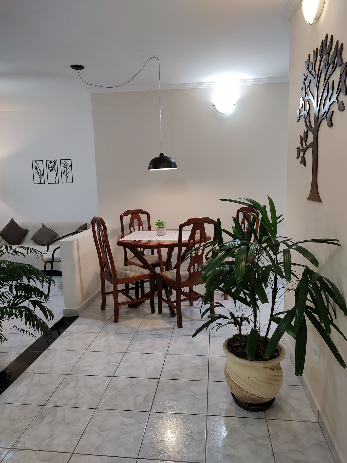 A dining area features a round table surrounded by four wooden chairs. A small potted plant sits at the center. Dim lighting is provided by a hanging pendant above, while a decorative wall piece and a planter with greenery add a fresh touch to the space.