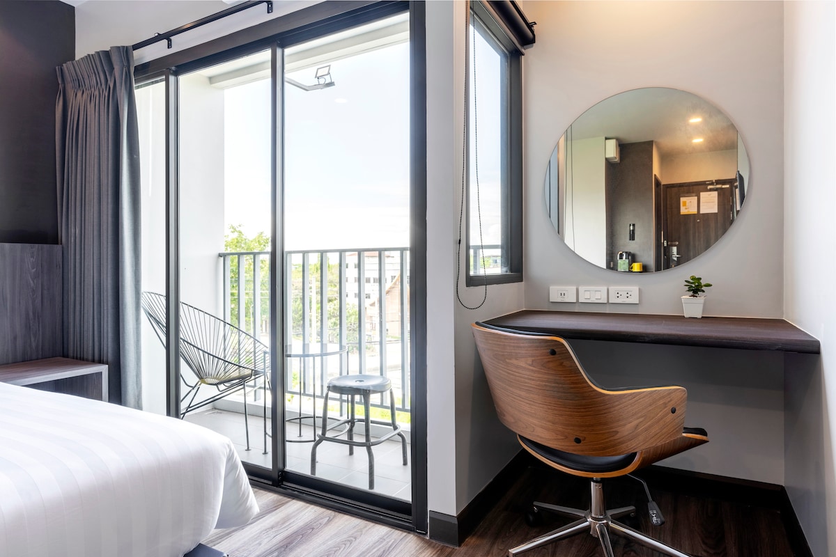 A bright bedroom area features a spacious bed beside a large sliding glass door leading to a balcony. A round mirror is mounted above a dark wood desk, complemented by a stylish chair. Natural light enhances the modern decor and serene ambiance.