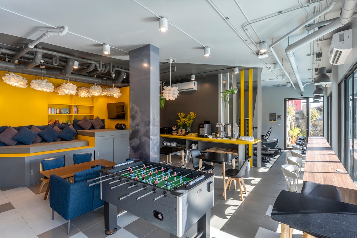 A welcoming co-living area features a foosball table at the center, surrounded by modern seating arrangements. Bright yellow accents enhance the space, complemented by plush cushions against the walls. Large windows allow natural light to flow in, contributing to a vibrant atmosphere.