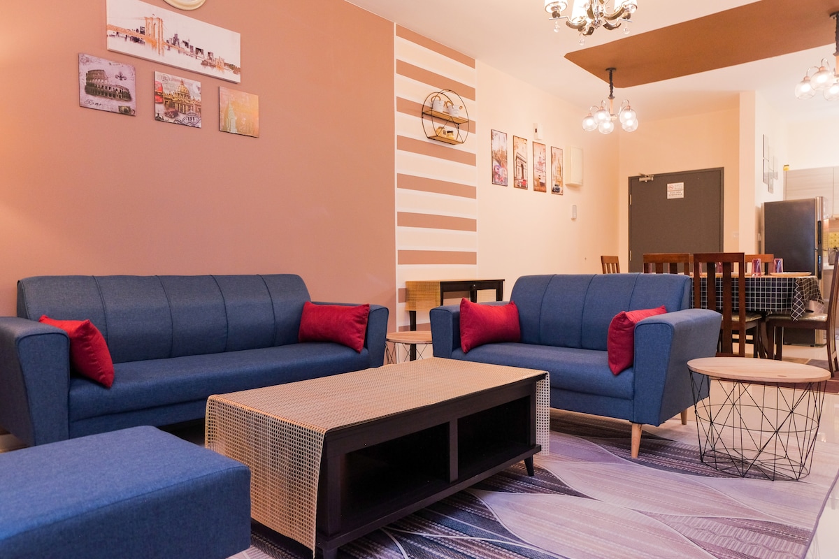 A modern living area showcases two blue sofas adorned with red cushions, complemented by a coffee table and a side table. Artwork and photographs line the wall, while a dining area with a table and chairs is visible in the background.
