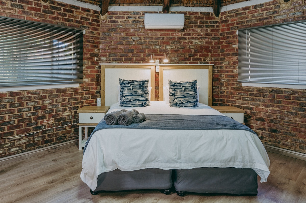 A cozy sleeping area features a spacious bed with crisp white linens and decorative pillows. The exposed brick walls add warmth, complemented by natural light from the large windows, and soft towels are neatly arranged at the foot of the bed.