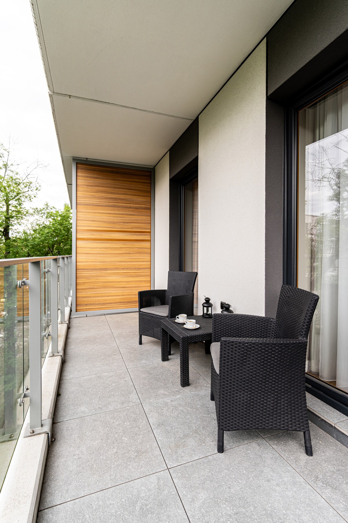 A private balcony is equipped with two black wicker chairs and a matching table. Small cups and a coffee pot are placed on the table. Large glass doors provide ample natural light and a view of surrounding greenery.