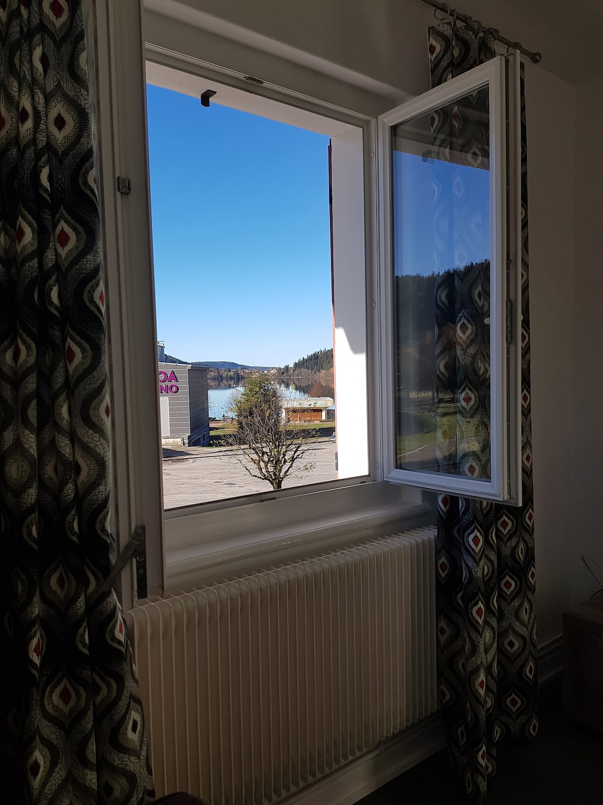 A white window is shown partially open, revealing a clear blue sky and a glimpse of the surrounding landscape. Decorative curtains with a geometric pattern frame the window, while a radiator is visible beneath it.