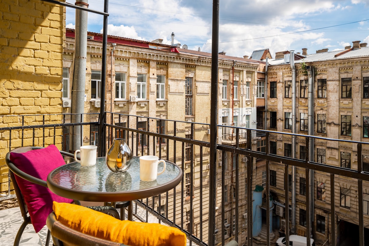 A balcony view reveals a charming courtyard, framed by historic building façades. A small round table is set with two mugs and a decorative bottle, complemented by colorful cushions on each side. The sky is partly cloudy, suggesting a pleasant atmosphere.