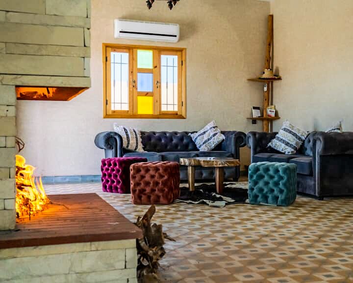 A cozy living area is arranged with plush sofas featuring colorful ottomans in shades of pink and teal. A wooden coffee table sits on a patterned tile floor, and a fireplace adds warmth to the space. Natural light filters through the windows.