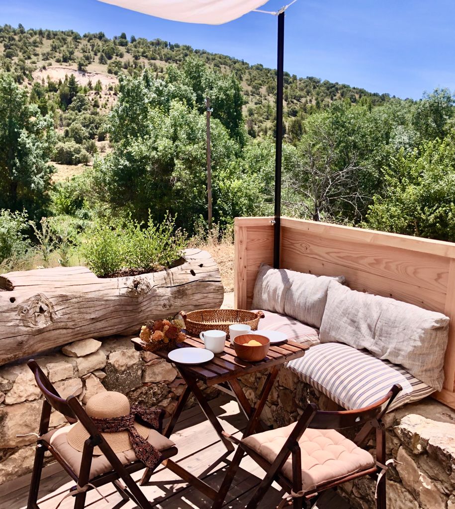 A cozy outdoor seating area features a small table set with dishes and a woven basket. Soft cushions are placed against a wooden backdrop, surrounded by greenery and scenic views of the hills, creating an inviting space for relaxation.