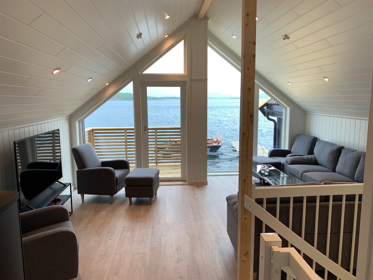 The airy living area features large windows that provide views of the water. Comfortable gray seating arrangements create a relaxed atmosphere. A sleek television is positioned for easy viewing. Natural light is abundant, enhancing the room's inviting and open feel.
