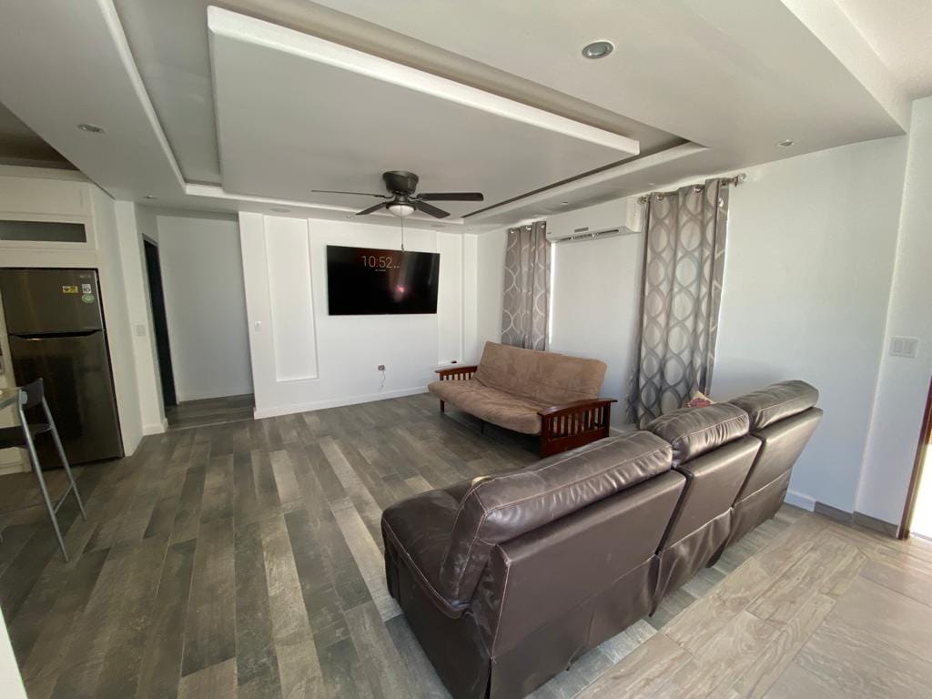The living area features a brown leather sofa facing a wall-mounted television. A light-colored futon sits nearby, complementing the neutral-toned walls. Large windows are adorned with sheer curtains, allowing natural light to fill the space, while a fan is positioned overhead.