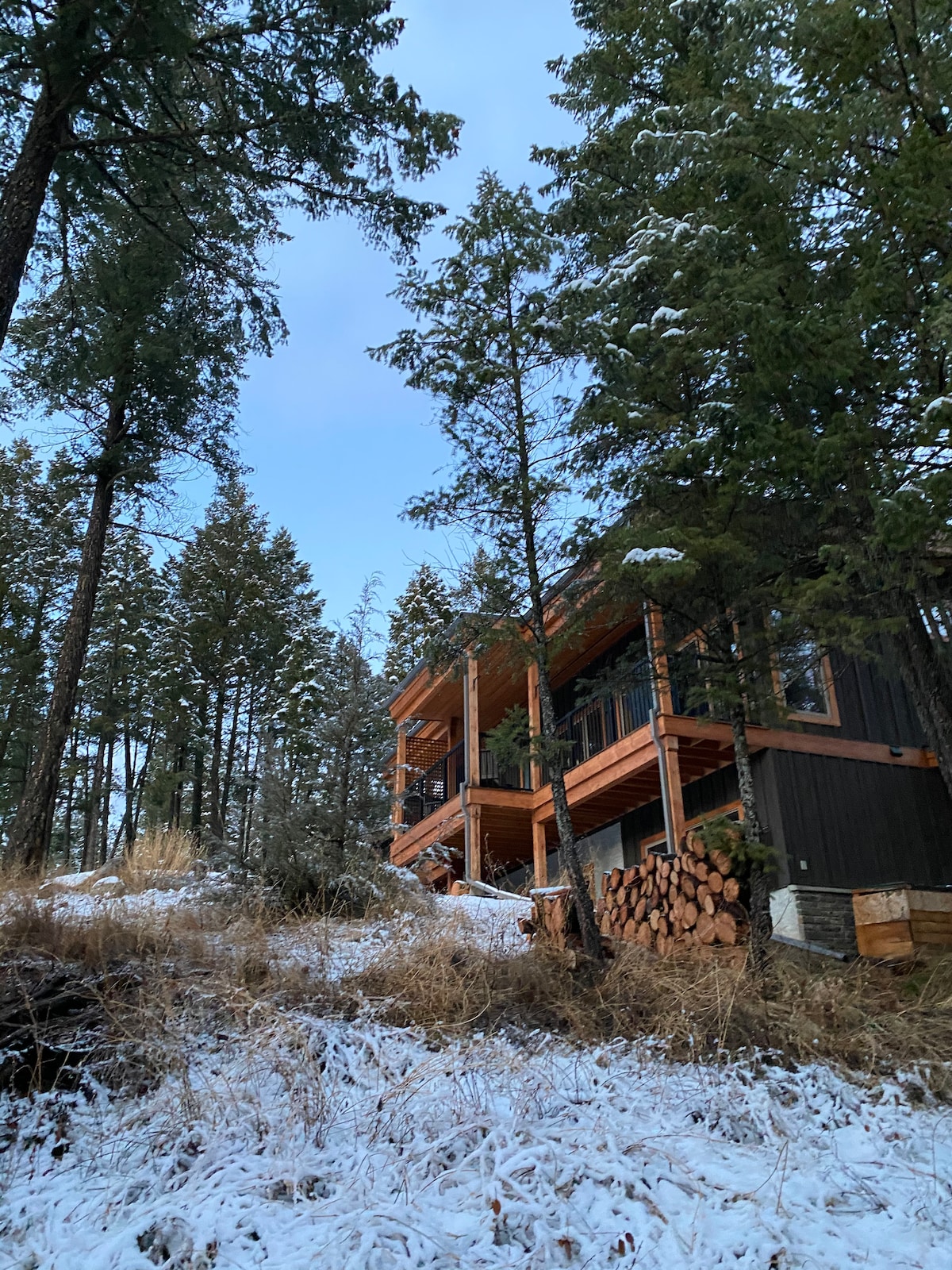A two-story cabin is surrounded by tall evergreen trees, with patches of snow covering the ground. Stacked logs are neatly arranged beside the cabin's exterior, showcasing a rustic charm. The structure features wooden balconies that overlook the natural landscape.