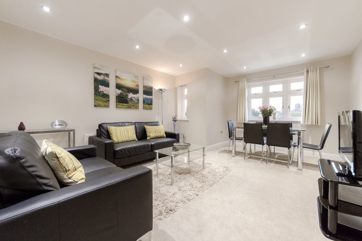 A spacious living area features comfortable leather sofas and a glass coffee table on a soft rug. A dining table for four is positioned near large windows, allowing natural light. Three landscape artworks adorn the wall, contributing to the modern decor.