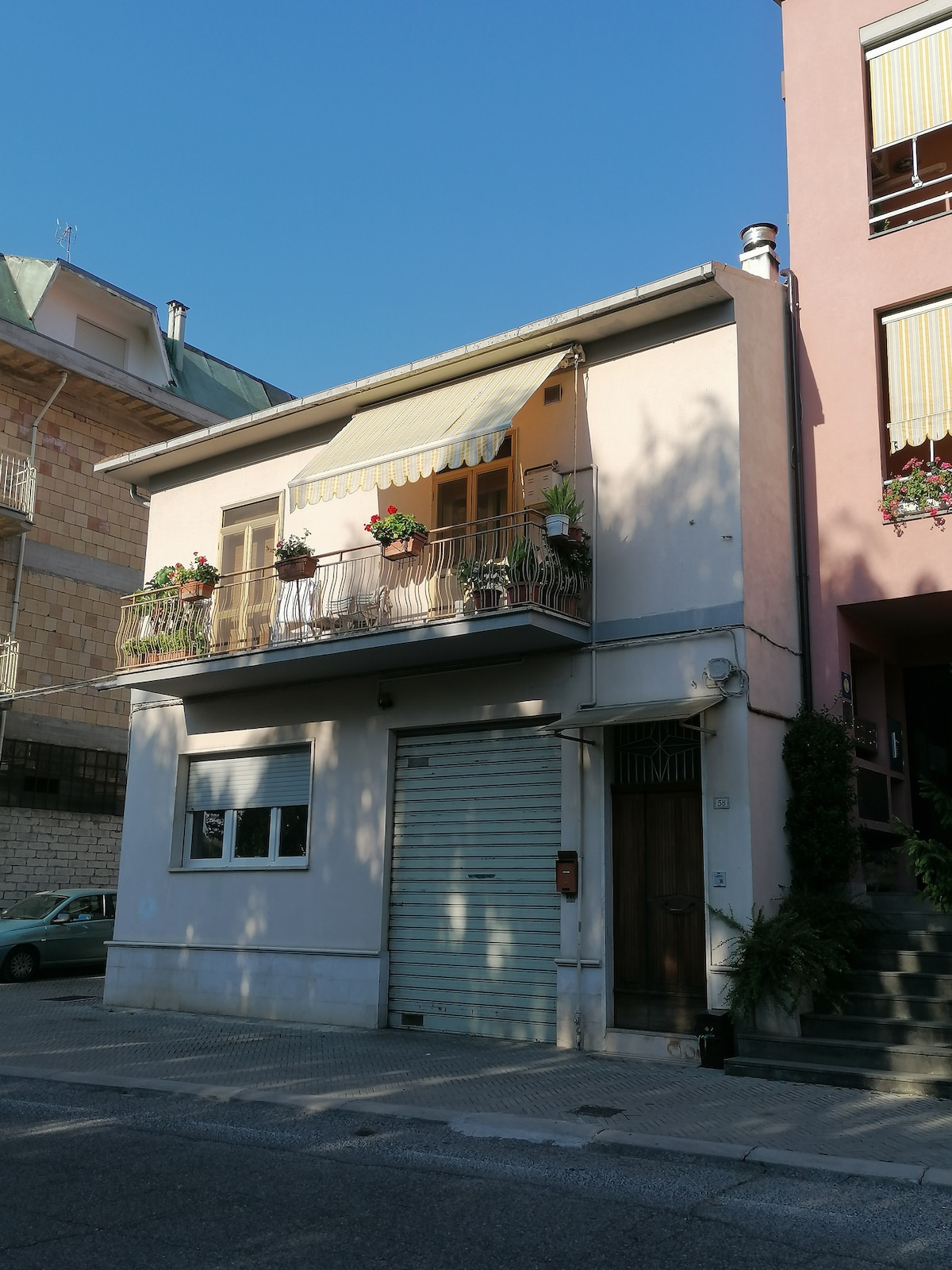 A charming two-story building features a light-colored facade with a balcony adorned with potted plants. The ground floor includes a garage door, while the entrance is marked by a wooden door and steps leading to the next level.