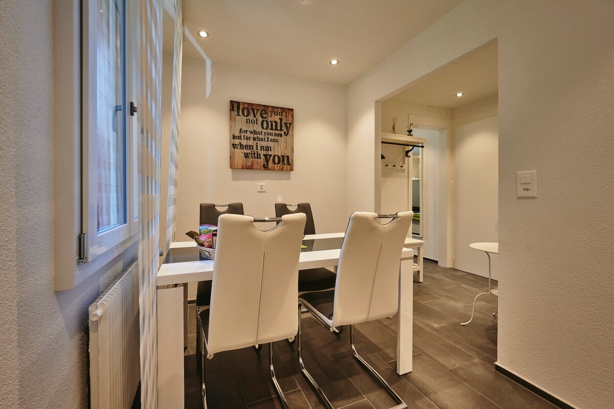 A modern dining area features a sleek white table surrounded by four white chairs. Soft lighting highlights the contemporary decor, including a framed wall art piece. Large windows allow natural light to brighten the space, contributing to an open and inviting atmosphere.