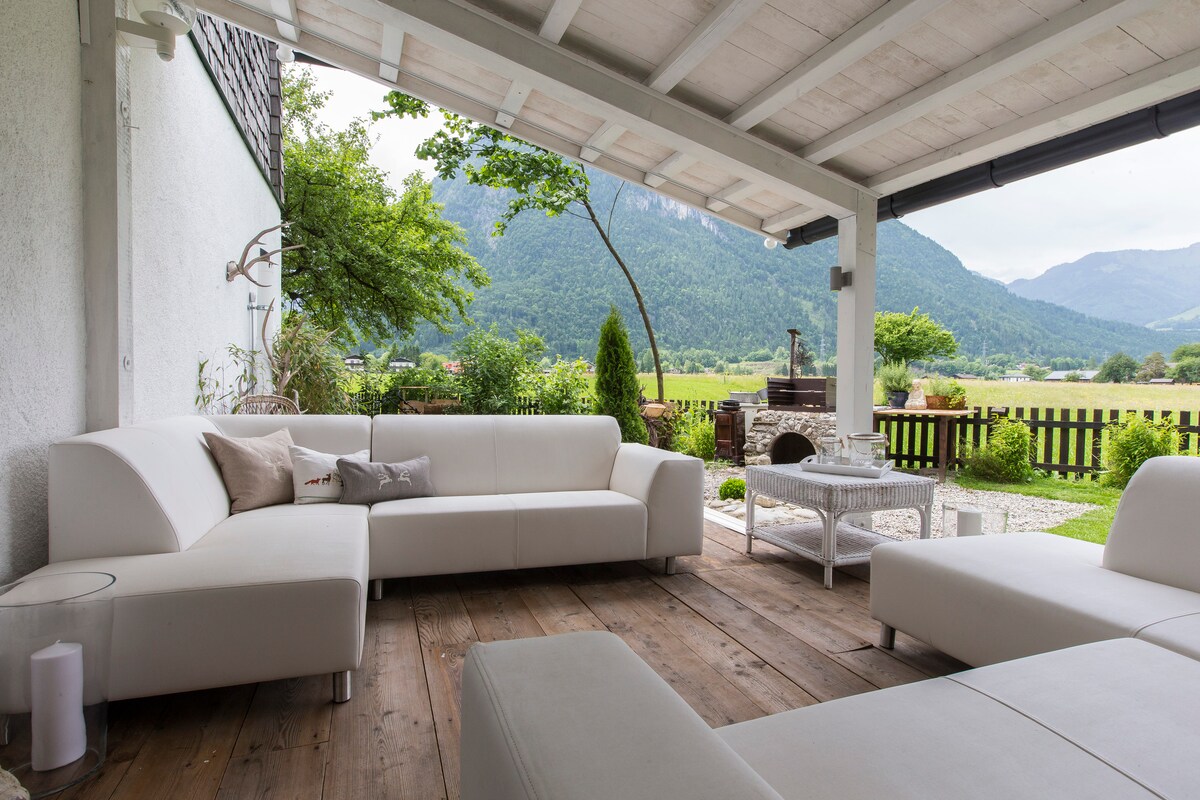 An open terrace features modern white lounge furniture arranged for relaxation. The wooden flooring contrasts with the sleek design, while a scenic view of green fields and mountains is visible in the background, creating a serene outdoor setting.
