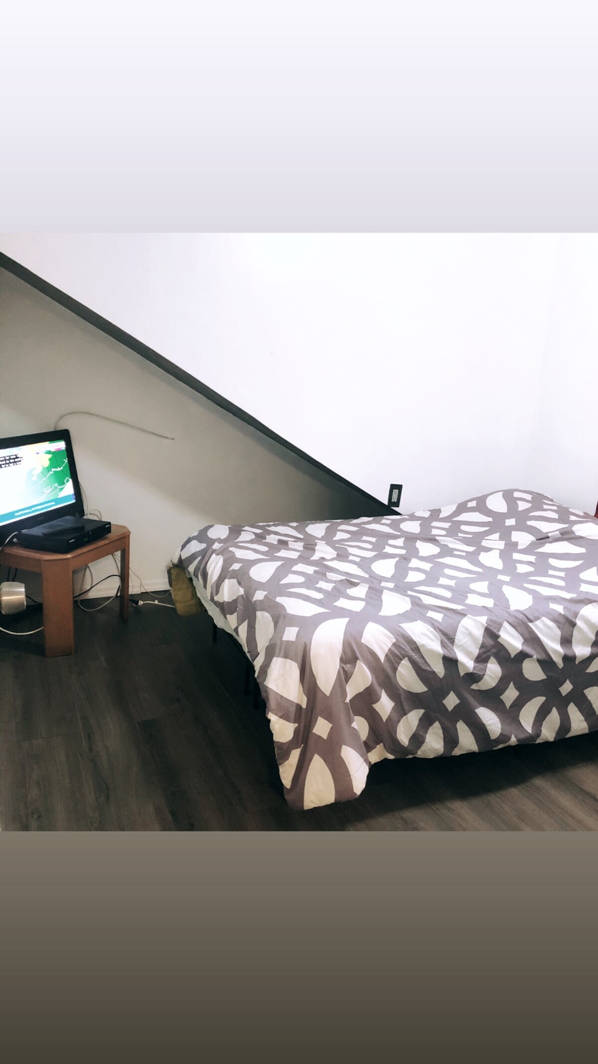 A cozy sleeping area features a full size bed covered with a patterned blanket. A small wooden table accompanies the bed, housing a television. The hardwood floor complements the minimalist design while the wall's slope adds architectural interest.