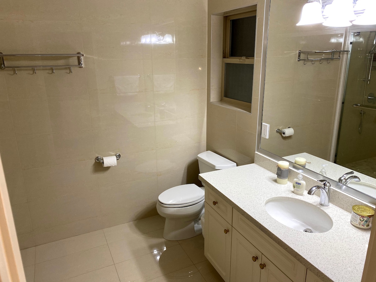 The ensuite bathroom features neutral-colored tiles and a clean design. A spacious vanity with a sink is accompanied by a large mirror above it. A toilet and a walk-in shower are also present. Towel hooks and toiletries are neatly arranged.