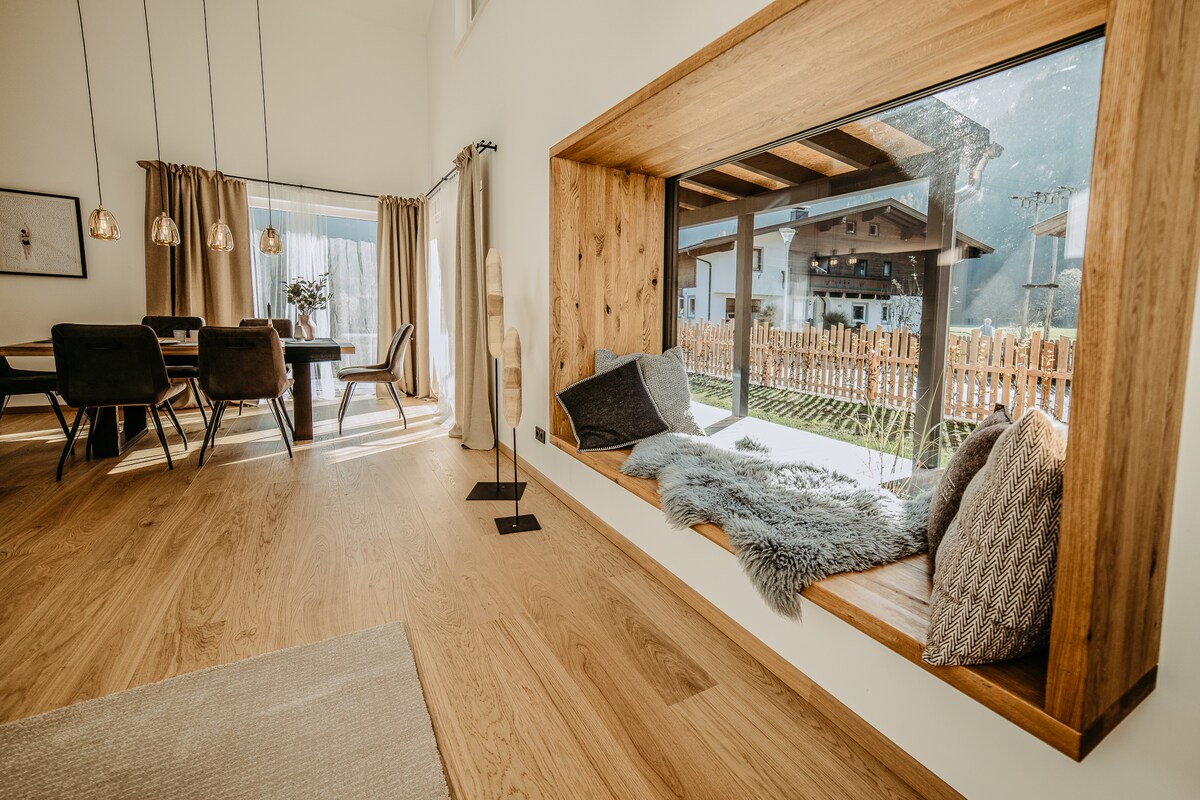 A cozy window nook is positioned next to a spacious living area, featuring soft cushions and a plush throw. The room is illuminated by natural light, with elegant dining arrangements visible in the background and large windows offering views of the garden and surrounding landscape.