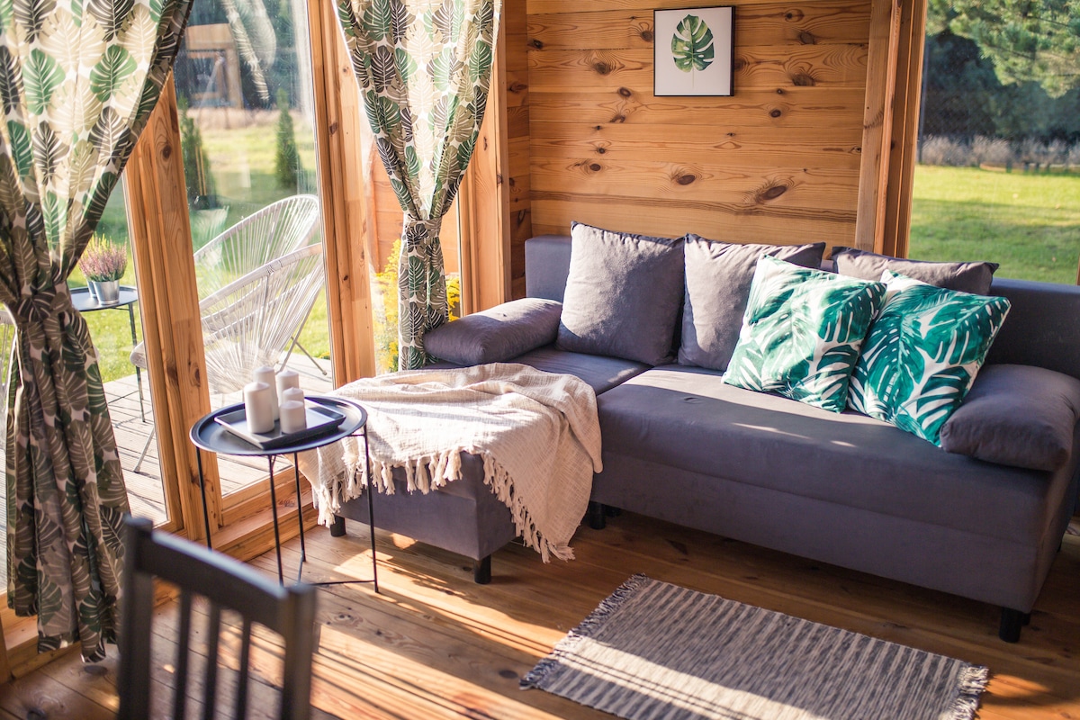 A welcoming seating area features a grey sofa adorned with textured cushions in green tones. Large windows allow natural light to fill the space, with soft curtains framing the outdoor view. A small table holds decorative items, and a woven rug adds warmth to the wooden floor.