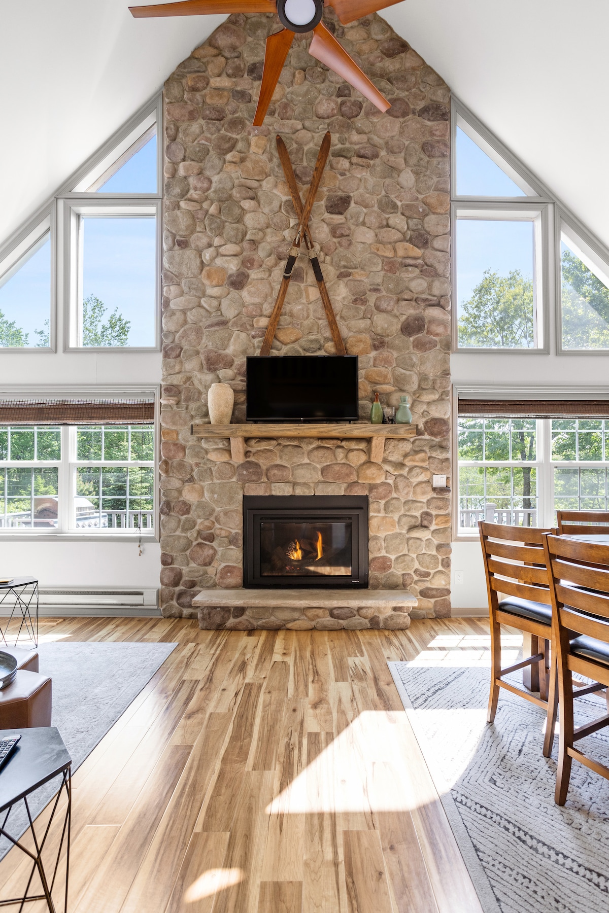 A spacious living area features a stone fireplace, complemented by light wooden flooring. Large windows allow natural light to fill the space, while a mounted TV is positioned above the fireplace. A dining table is visible, offering seating for multiple guests.