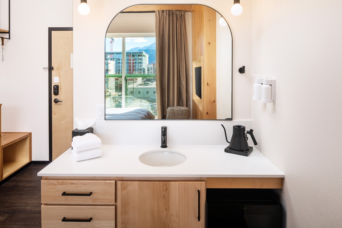A modern vanity sink area is presented with a large mirror and stylish lighting. A black kettle and white towels are neatly arranged, while a small refrigerator is visible beneath the counter. The reflection of a mountain view enhances the space's overall appeal.