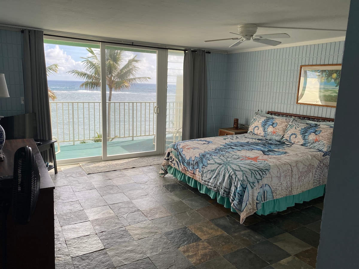 A spacious bedroom features a large bed with a colorful quilt, complemented by natural stone flooring. Sliding glass doors open to reveal ocean views, framed by light gray curtains. A wooden nightstand and a dresser are positioned beside the bed, enhancing the room's functionality.