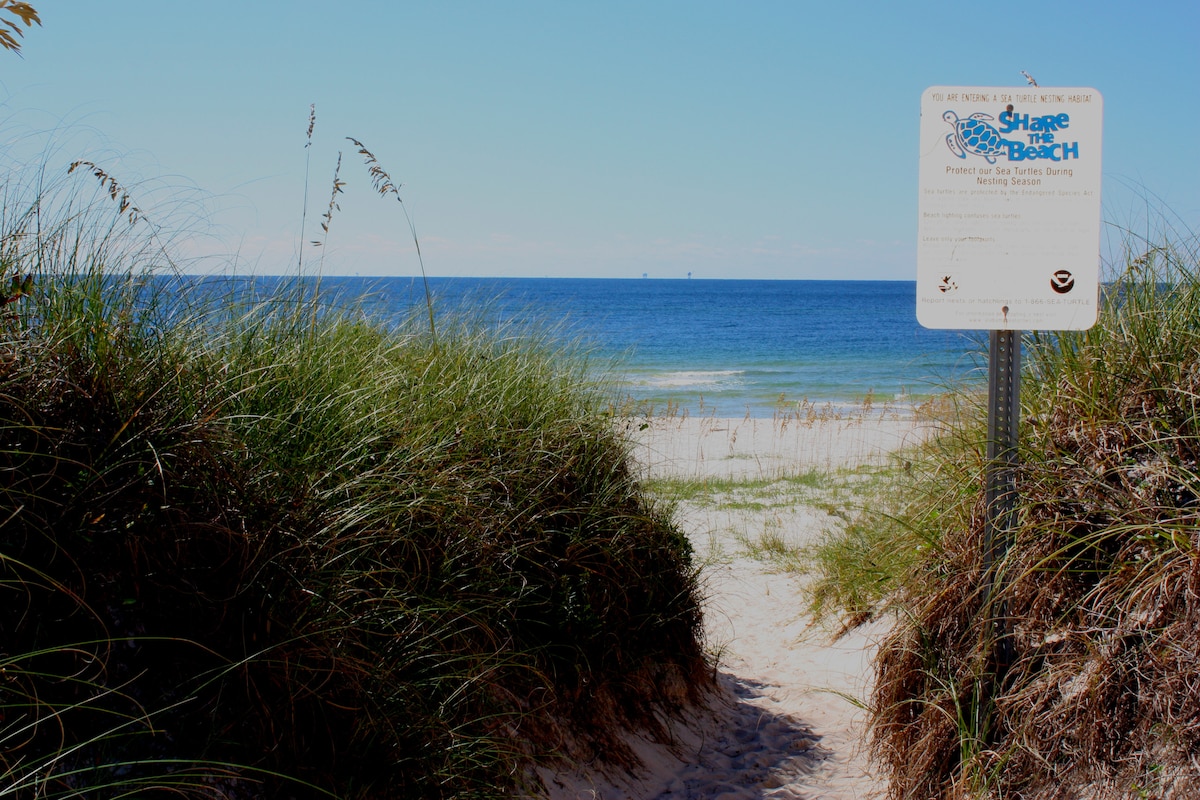 A sandy pathway leads through tall grass to a calm beach area. A sign marking the beach features information about the private access. The tranquil blue waters of the Gulf of Mexico are visible in the background, complemented by a clear sky.