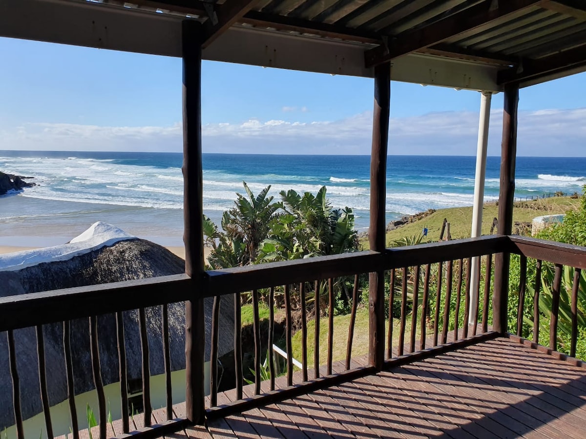 A wooden deck showcases sweeping ocean views, with gentle waves rolling onto the sandy beach. Lush greenery is visible in the foreground, complementing the coastal landscape. The clear blue sky adds to the serene environment, creating an inviting space for relaxation.
