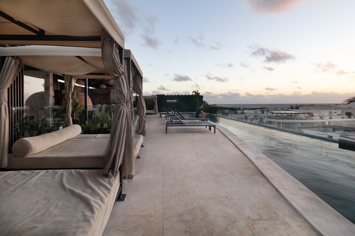 A serene rooftop space features comfortable cabanas alongside loungers, providing a tranquil setting overlooking the horizon. The sleek infinity pool extends into the distance, reflecting the soft evening light and creating a peaceful ambiance. Lush greenery adds a touch of nature to the modern design.