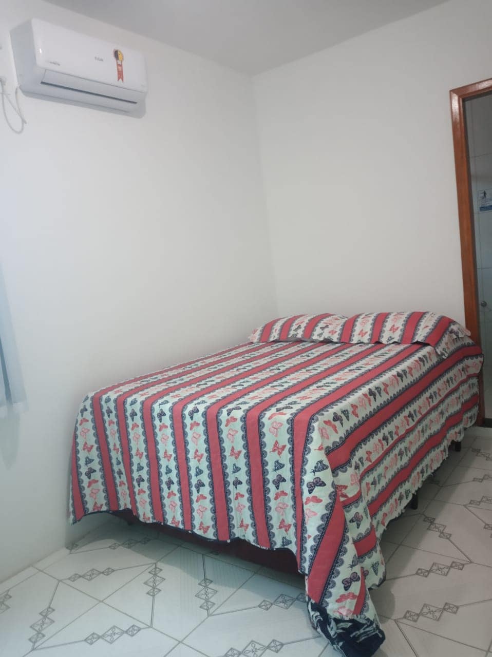 A sleeping area features a spacious bed with a patterned blanket. The light-colored walls create a bright atmosphere, while an air conditioning unit is mounted above. The floor displays a tiled design, and a door leads to a private bathroom.
