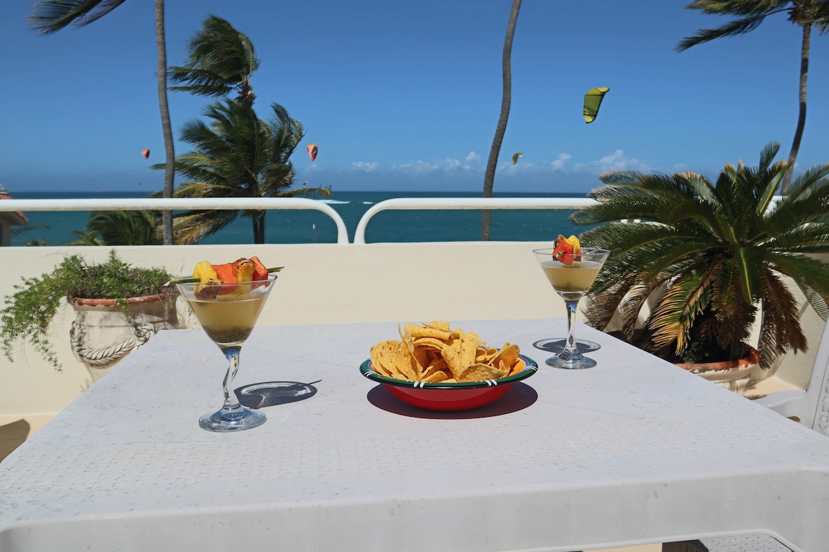 A panoramic view of the ocean is featured from a rooftop terrace, complemented by two glasses of fruit cocktails and a bowl of snacks on a white table. Palm trees sway gently in the breeze, while colorful kites can be seen in the distance against a clear blue sky.