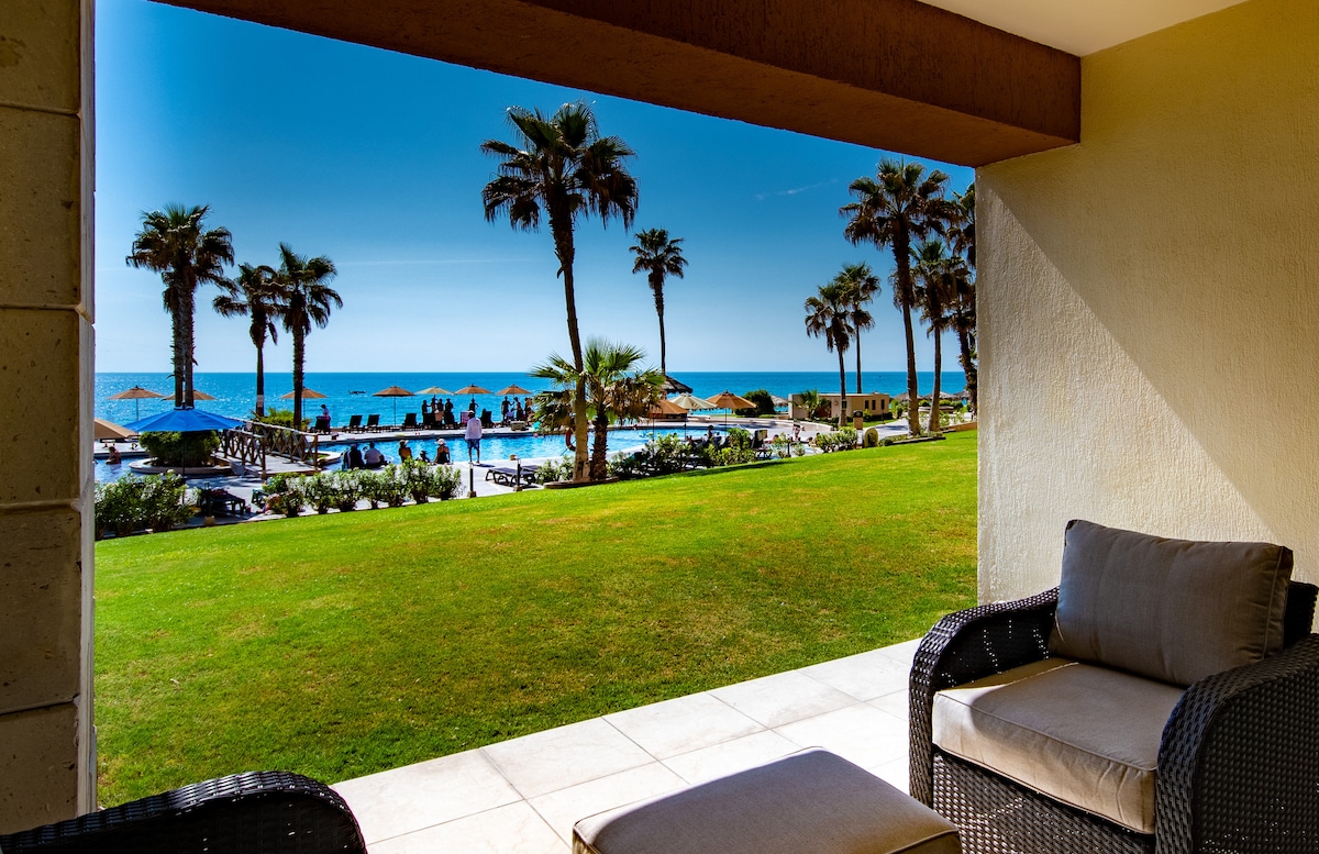 A serene outdoor patio is showcased, featuring comfortable seating that overlooks the lush green lawn, pool area, and the ocean beyond. Palm trees frame the view, creating a relaxing atmosphere. The bright blue sky complements the inviting landscape.