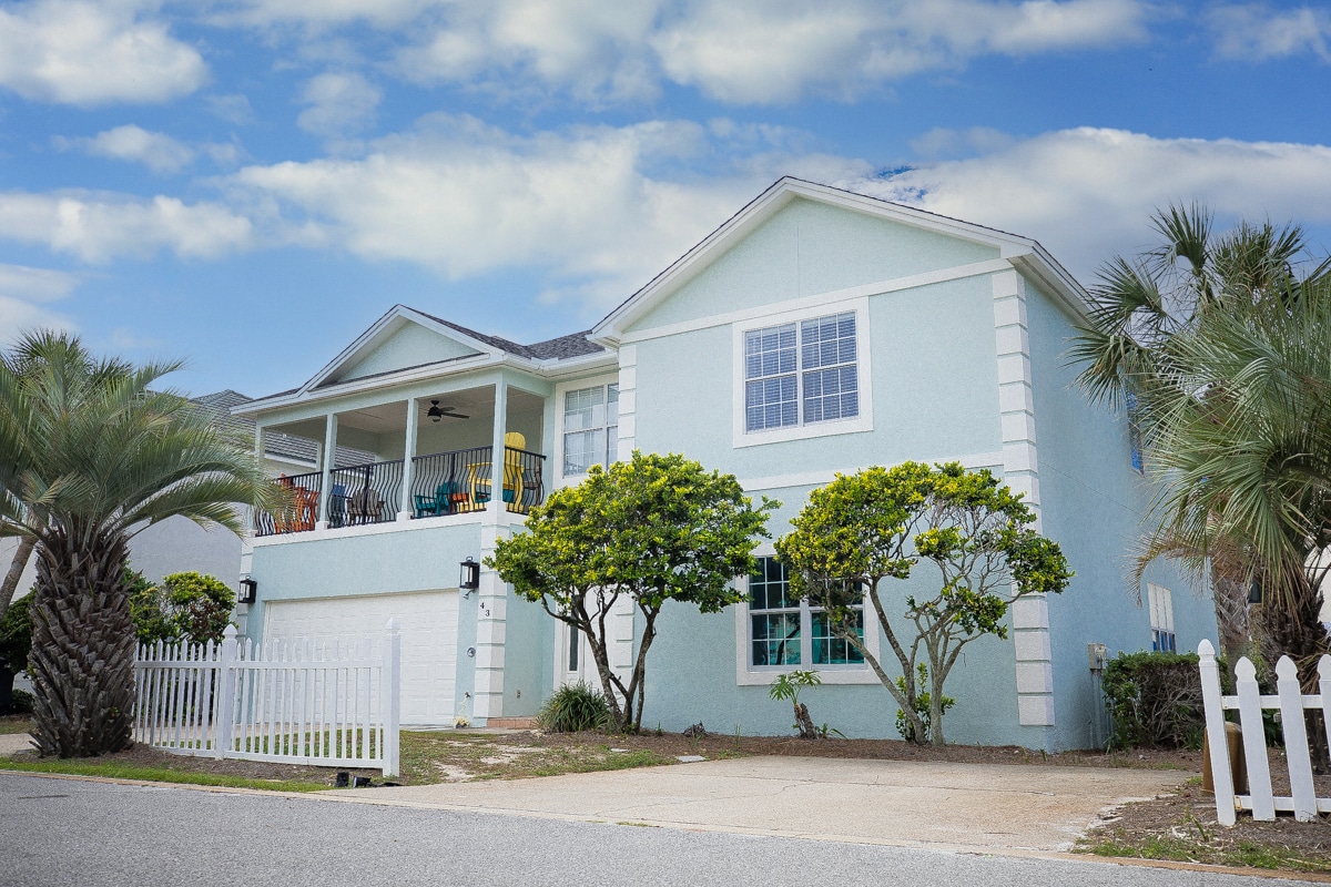 A light blue, two-story house is surrounded by palm trees and shrubbery. A white picket fence outlines the front yard, adding to the welcoming aesthetic. A covered balcony is visible on the second floor, furnished with colorful chairs, offering a spot for relaxation.