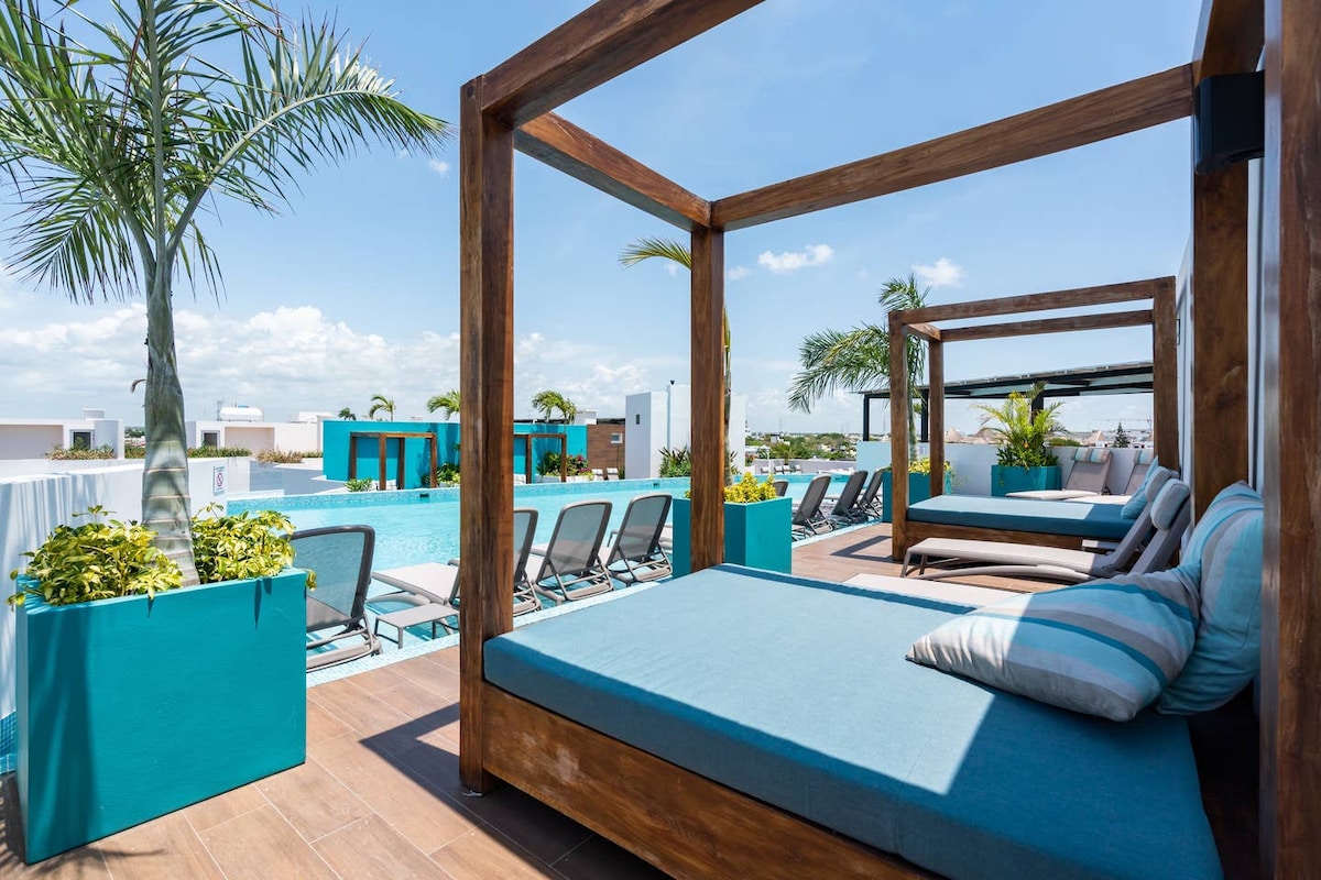 A rooftop terrace features several wooden cabanas with cushioned lounge areas, surrounded by vibrant potted plants and palm trees. A swimming pool is visible in the background, with a row of sun loungers arranged nearby, creating a space for relaxation.