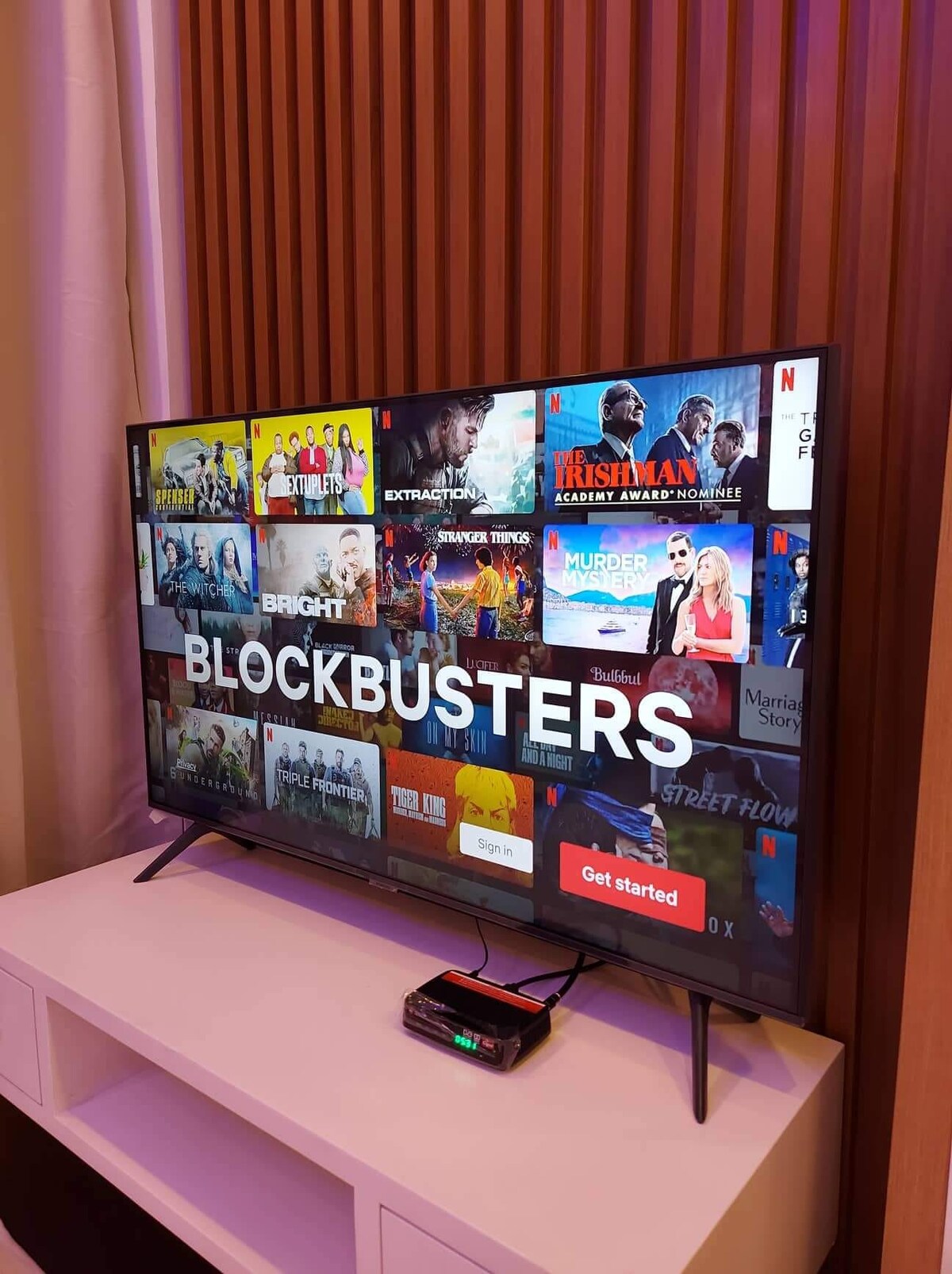A smart television displays a selection of movies and shows, featuring a prominent 'Blockbusters' title. The TV is positioned on a white entertainment unit with a small device located underneath it. Soft lighting and a wooden backdrop enhance the viewing area.
