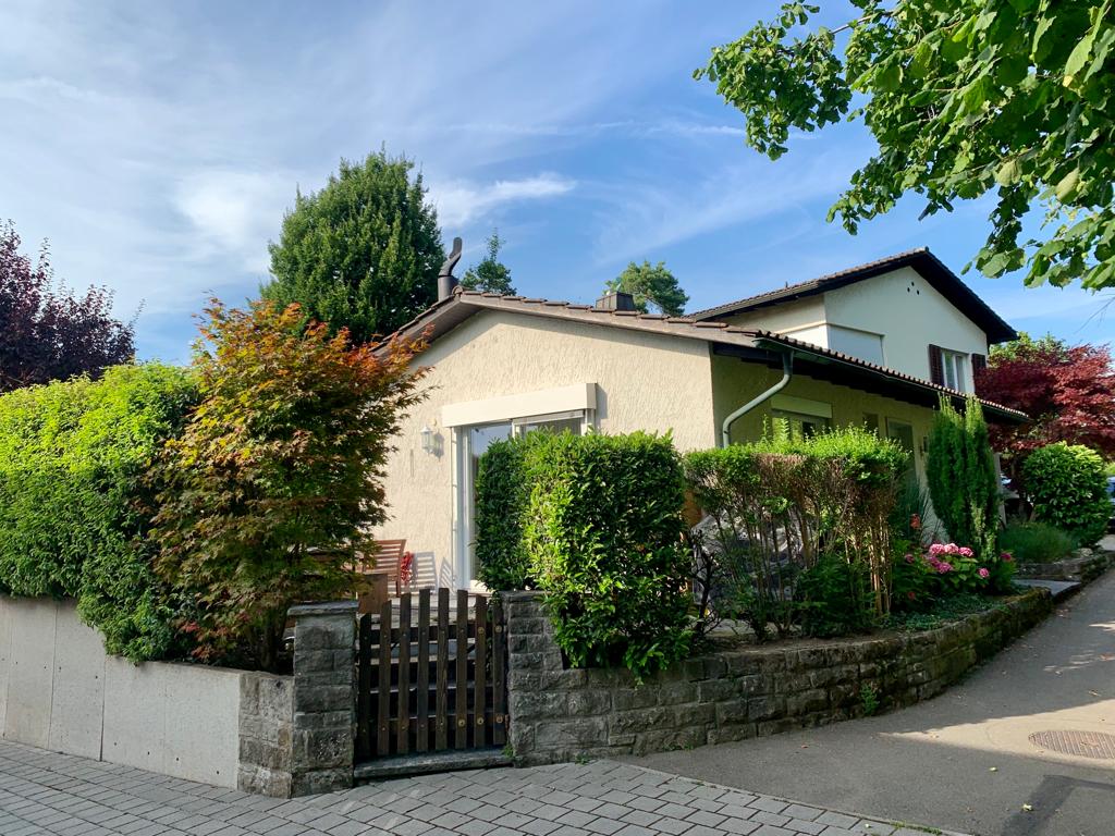 The exterior of a welcoming apartment is framed by lush greenery and colorful plants. A wooden gate offers access to the property, which features a neatly landscaped yard. The facade presents a blend of light-colored walls and a darker roof under a clear blue sky.