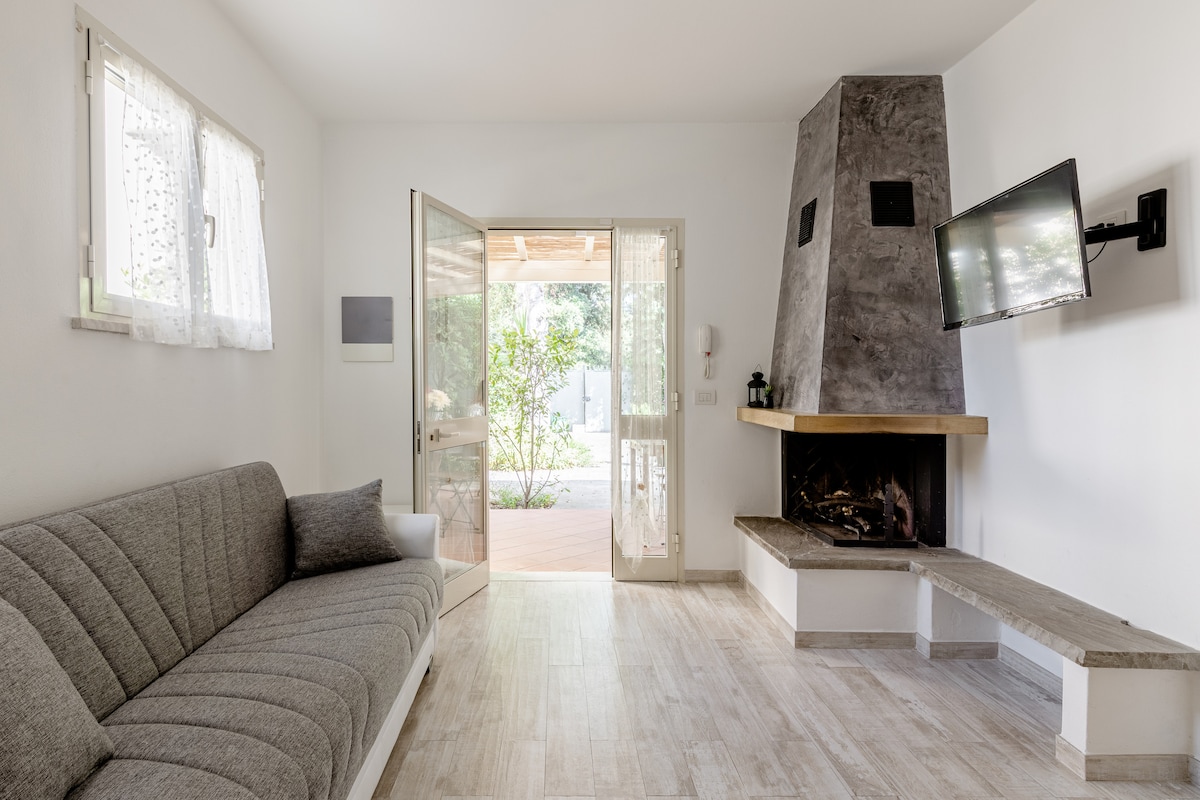 A bright living room features a comfortable gray sofa positioned against a wall. A stone fireplace is built into the corner, with a flat-screen television mounted nearby. Double doors open to a patio, inviting natural light and views of the surrounding greenery.