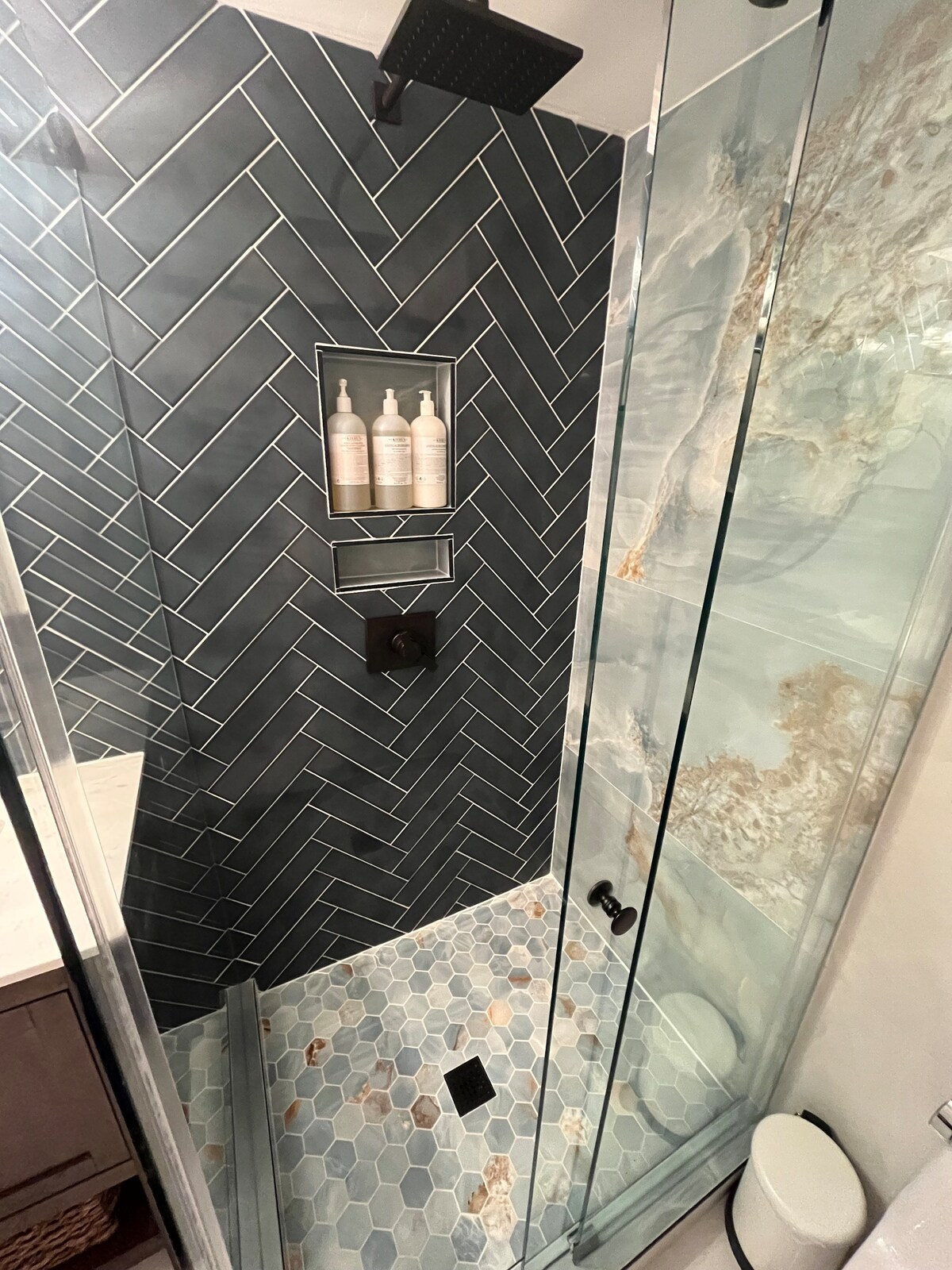 A modern shower area features sleek glass walls and a textured tile design in herringbone pattern. Shelves hold neatly arranged toiletries. The flooring is composed of hexagonal tiles with a subtle color variation, highlighting the contemporary aesthetic.