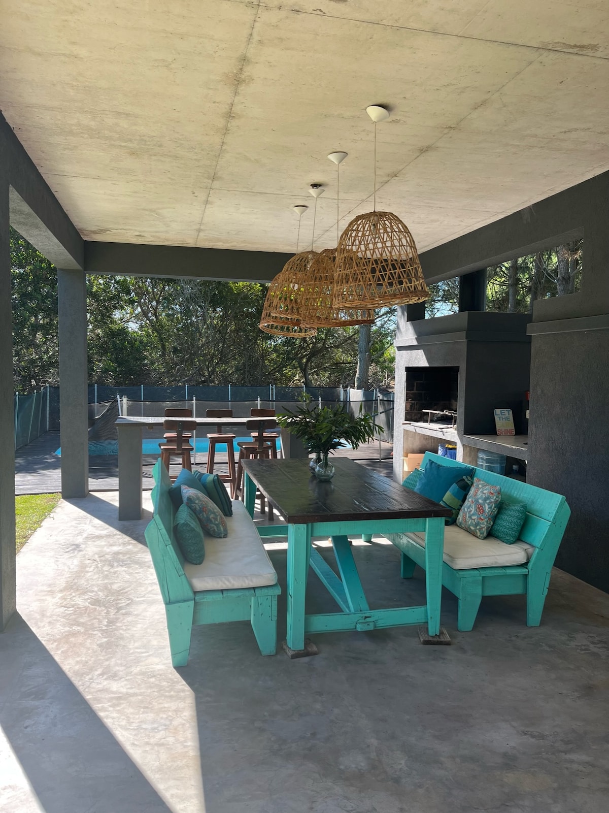 An outdoor dining area is featured under a covered porch, with a wooden table and bench seating. Two large pendant lights hang overhead. To the side, a barbecue grill is positioned next to a kitchenette. A turquoise pool is visible in the background, surrounded by greenery.