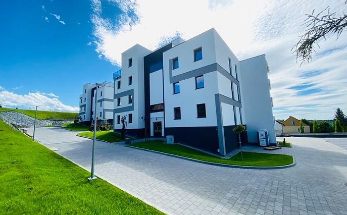 A modern, multi-story building features a sleek design with large windows and contrasting color blocks. The surrounding area is landscaped with green grass and a paved pathway, under a bright blue sky with scattered clouds.
