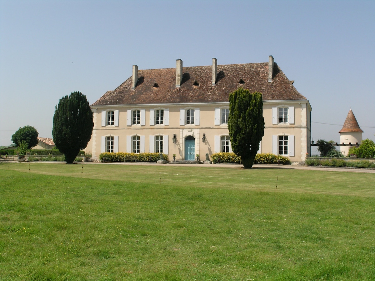 A grand château is depicted with a light beige façade and a sloped roof. Two tall trees frame the entrance, while neatly trimmed hedges are positioned in front. A round tower is seen in the background, set against a clear blue sky.