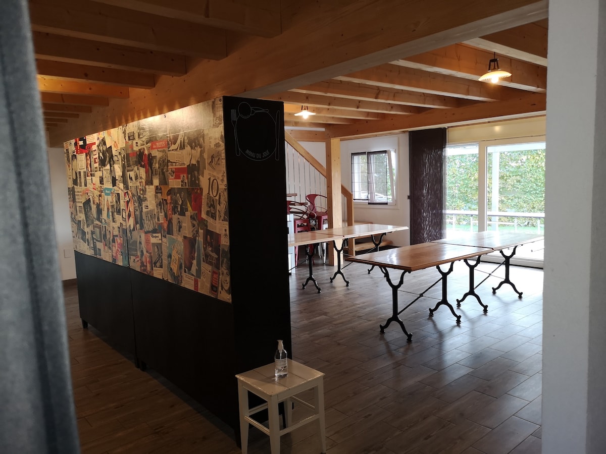 An open meeting space is featured, showcasing a large black partition adorned with various images and notes. Several folding tables are arranged for group activities, and large windows allow natural light to fill the area, creating a bright and airy environment.
