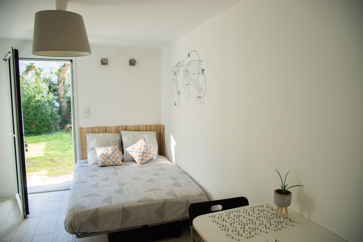 A cozy studio space features a comfortable bed with patterned decorative cushions. A small dining table is nestled beside it. Natural light pours in from the open doors, leading to a green outdoor area. A simple world map decorates the wall, enhancing the ambiance.