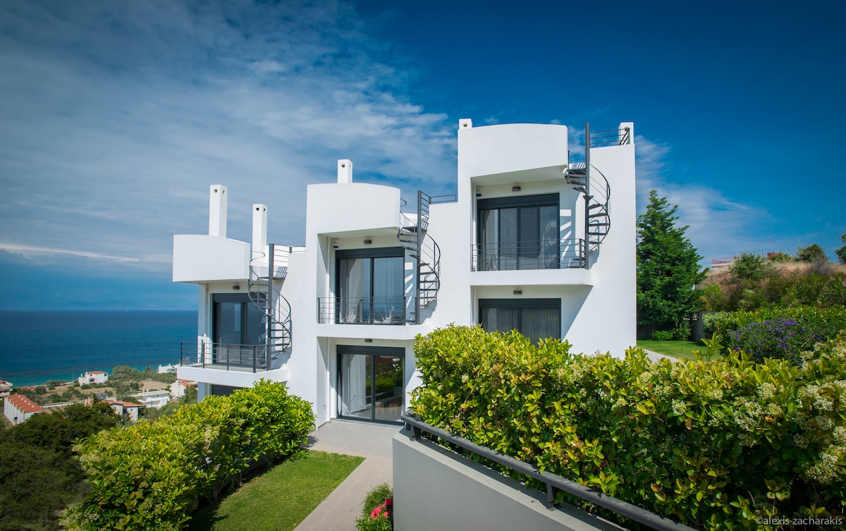The modern building is positioned in a scenic location overlooking the sea. It features three levels with a sleek white exterior and large glass windows. A landscaped garden with greenery surrounds the entrance, complemented by a pathway leading up to the front door.